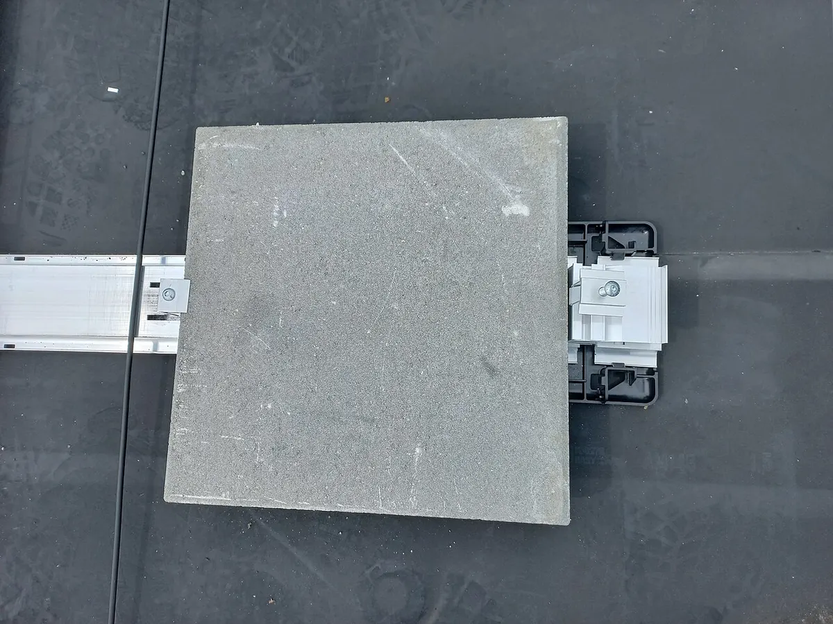 Widok z góry na kwadratową betonową płytę położoną jako balast na aluminiowej szynie montażowej i plastikowej stopie, na czarnej membranie dachu płaskiego