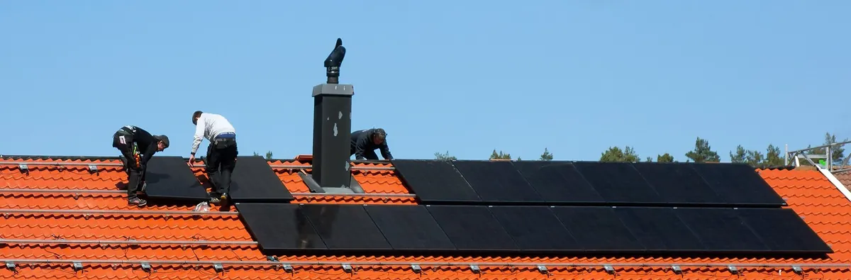 Trzech instalatorów na dachu krytym dachówką montuje w rzędach czarne moduły fotowoltaiczne wokół komina