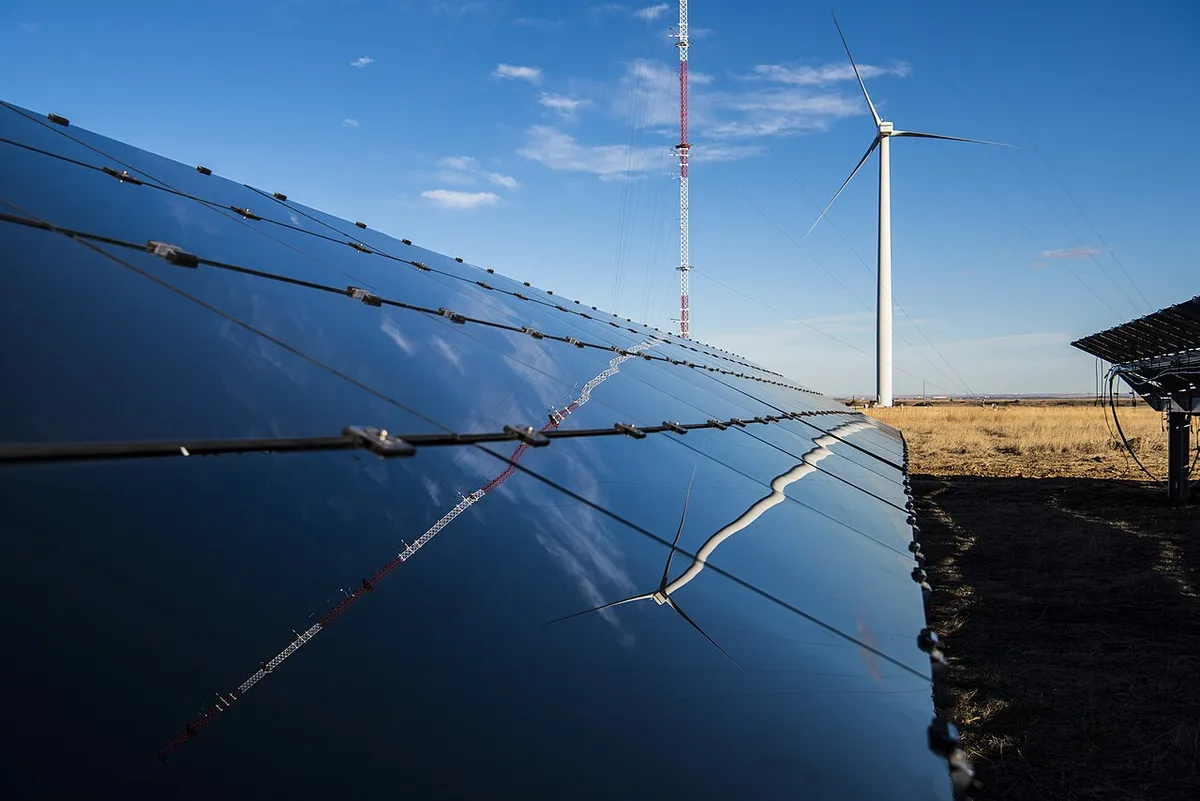 Rząd jednolicie ciemnych, bezramowych modułów cienkowarstwowych CdTe z odbiciem nieba, w tle turbina wiatrowa i maszt pomiarowy na stepie
