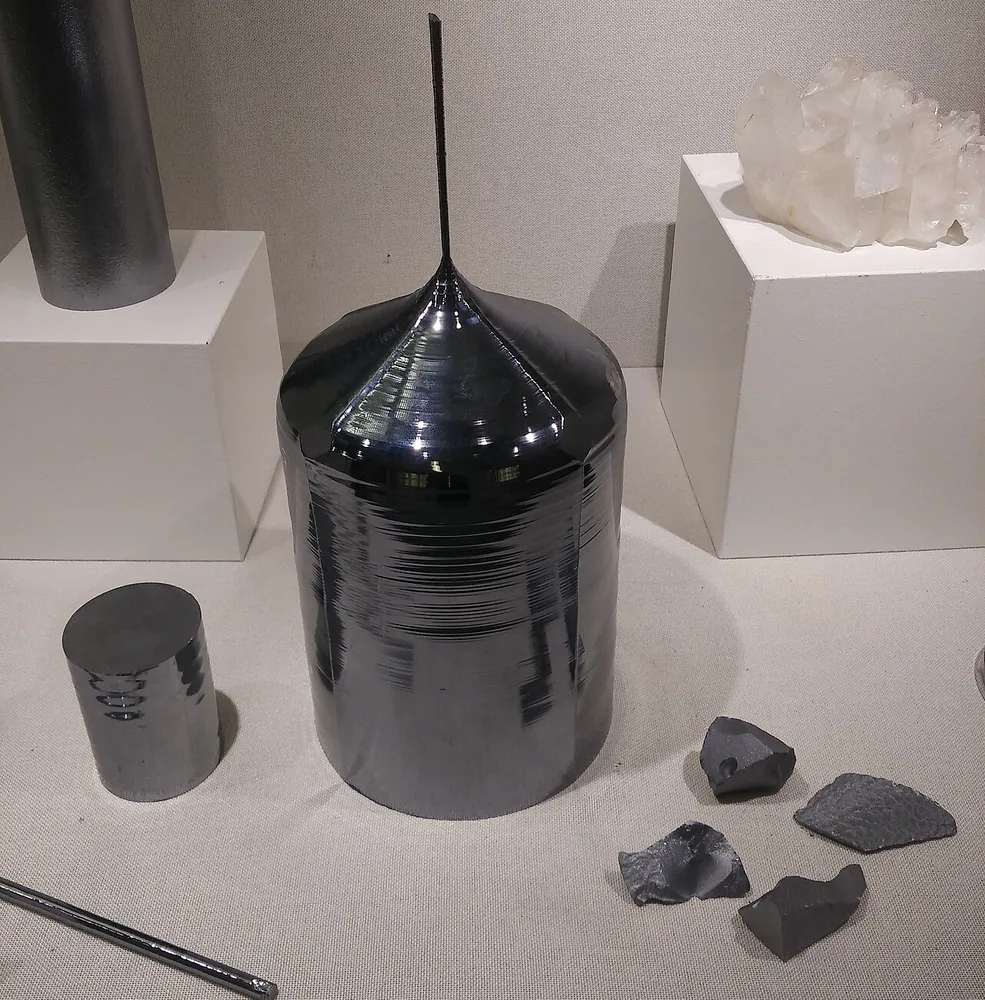 Ciemny, błyszczący cylindryczny wlewek krzemu monokrystalicznego ze stożkowym czubkiem i cienkim prętem zarodkowym na muzealnym postumencie, obok mniejszy walec krzemu i okruchy surowca