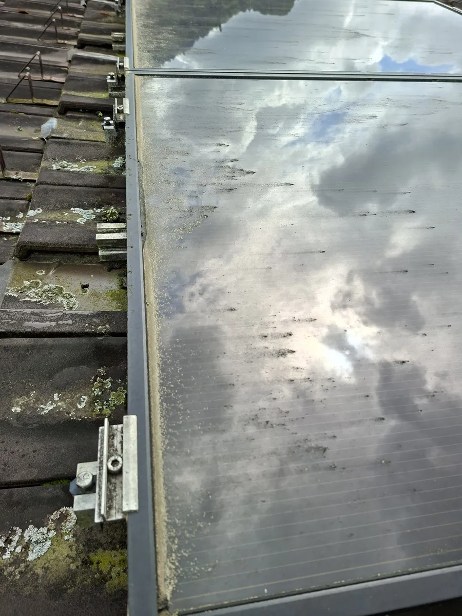 Zbliżenie cienkowarstwowego modułu fotowoltaicznego na dachu z dachówki, z pasem brudu i pyłku osadzonym wzdłuż dolnej krawędzi ramy i ciemnymi zaciekami na szybie