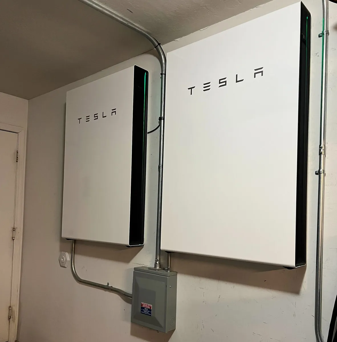 Dwa białe domowe magazyny energii Tesla Powerwall 3 zawieszone na ścianie w pomieszczeniu technicznym, pod nimi szara skrzynka rozłącznika