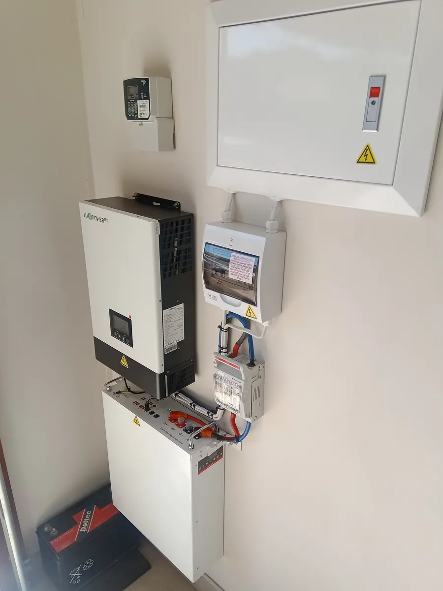 Falownik hybrydowy 5 kW na ścianie nad białym magazynem litowym 5,8 kWh, obok rozdzielnica z zabezpieczeniami