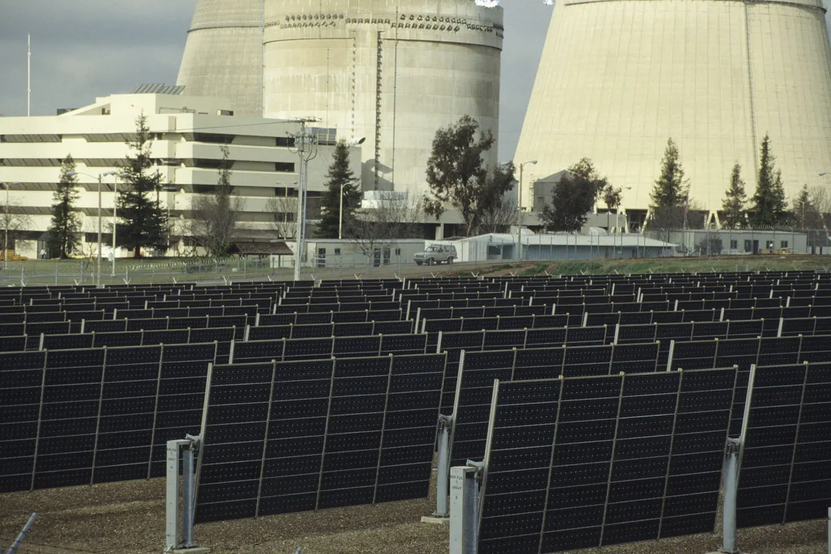 Rzędy ciemnych modułów fotowoltaicznych na jednoosiowych trackerach przed chłodniami kominowymi elektrowni Rancho Seco w Kalifornii